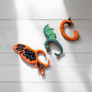 以下が含まれることがあります： 蝶、ドラゴン、巻き付いたタツノオトシゴの形をしたオレンジと緑のプラスチックのおもちゃ3つ。おもちゃは白い木製の表面の上に置かれています