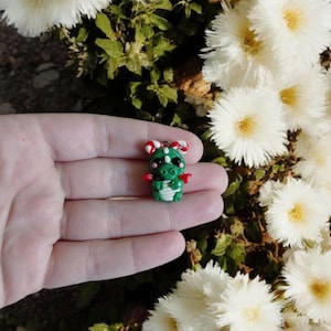 May include: A small, green dragon figurine with red wings and candy cane horns. The dragon has white accents and black eyes. The figurine is held in a hand, with white flowers and green foliage in the background.