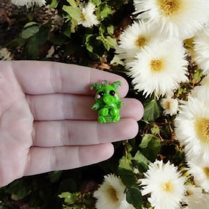 Op de afbeelding: Een kleine groene draakfiguur met roze wangen en kleine vleugels. De draak heeft een lus aan de bovenkant om op te hangen. De achtergrond toont witte bloemen en groen gebladerte.