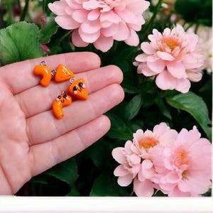 May include: Three orange clay charms in the shape of a crescent moon, a triangle, and a pizza slice. The pizza slice has a smiley face with two pink dots for eyes.