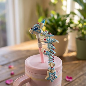 Puede incluir: Un vaso rosa claro con una pajita que presenta un adorno de dragón articulado y esculpido. El dragón es blanco con patrones de escamas azul claro, pequeños detalles rojos y grandes ojos azules. La pajita termina en una estrella blanca.