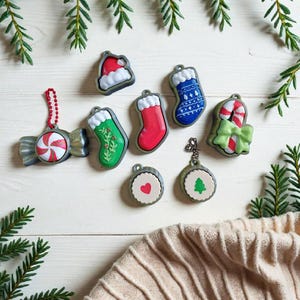 Puede incluir: Una colección de adornos navideños, que incluyen un gorro de Papá Noel, calcetines, un bastón de caramelo y un caramelo envuelto. Los adornos son rojos, verdes, azules y blancos, con un borde plateado. Están dispuestos sobre una superficie de madera blanca con ramas de pino.