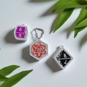 Pode incluir: Três chaveiros em forma de dados, brancos com detalhes em preto, vermelho e roxo. Os dados mostram números e são projetados para jogos de RPG de mesa. Os chaveiros estão em uma superfície branca com folhas verdes.