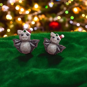 May include: Two gray bat ornaments with red and white Santa hats. The bats are hanging from a green velvet surface.