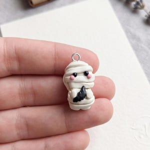 May include: A handmade mummy charm made of white clay, wrapped in bandages, with black eyes and a black object. The charm has pink cheeks and a silver loop for hanging. A small, antique book is in the background.