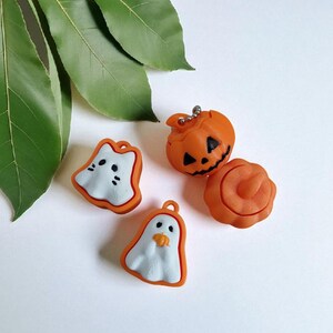 K&ouml;nnte beinhalten: Vier Halloween-Anh&auml;nger in Orange, Wei&szlig; und Schwarz. Die Anh&auml;nger umfassen zwei Geister, einen geschnitzten K&uuml;rbis und eine K&uuml;rbisform. Die Anh&auml;nger liegen auf einer wei&szlig;en Oberfl&auml;che mit gr&uuml;nen Bl&auml;ttern im Hintergrund.