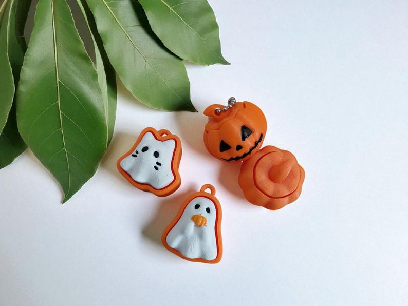 K&ouml;nnte beinhalten: Vier Halloween-Anh&auml;nger in Orange, Wei&szlig; und Schwarz. Die Anh&auml;nger umfassen zwei Geister, einen geschnitzten K&uuml;rbis und eine K&uuml;rbisform. Die Anh&auml;nger liegen auf einer wei&szlig;en Oberfl&auml;che mit gr&uuml;nen Bl&auml;ttern im Hintergrund.