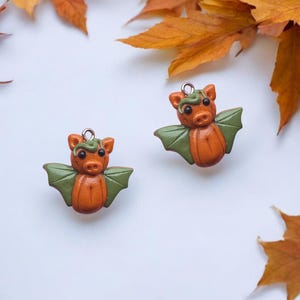 May include: Two orange and green bat-shaped charms with silver-colored loops for hanging. The bats have green wings and a green vine-like detail on their heads. The background is white with orange autumn leaves.