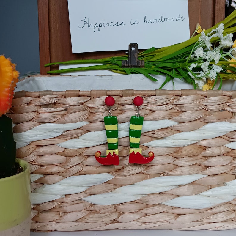 May include: A pair of elf leg earrings with red boots, green and yellow striped legs, and red tops. The earrings are hanging from a woven basket.