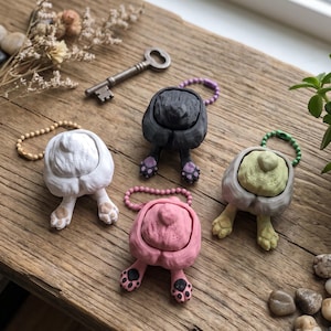 May include: Four animal-themed earbud cases in white, black, pink, and grey, each featuring a sculpted rear end with visible paws and a tail. Each case includes a small colored ball chain attachment for hanging, displayed as a set of four individual units.