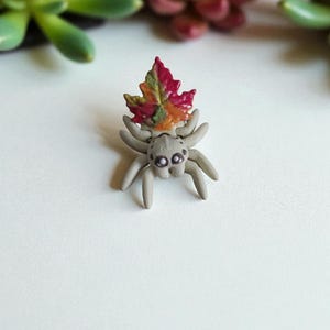 Könnte beinhalten: Handgefertigte, graue Spinnenfigur mit einem bunten Herbstblatt auf dem Kopf. Das Blatt zeigt Farbtöne von Rot, Orange und Grün. Die Spinne hat mehrere Beine und kleine, detaillierte Augen. Dieses einzigartige Stück ist wahrscheinlich ein Dekorationsartikel oder ein Sammlerstück.
