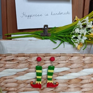 May include: A pair of elf leg earrings with red shoes, green and yellow striped legs, and gold bells. The earrings are displayed in a wicker basket.