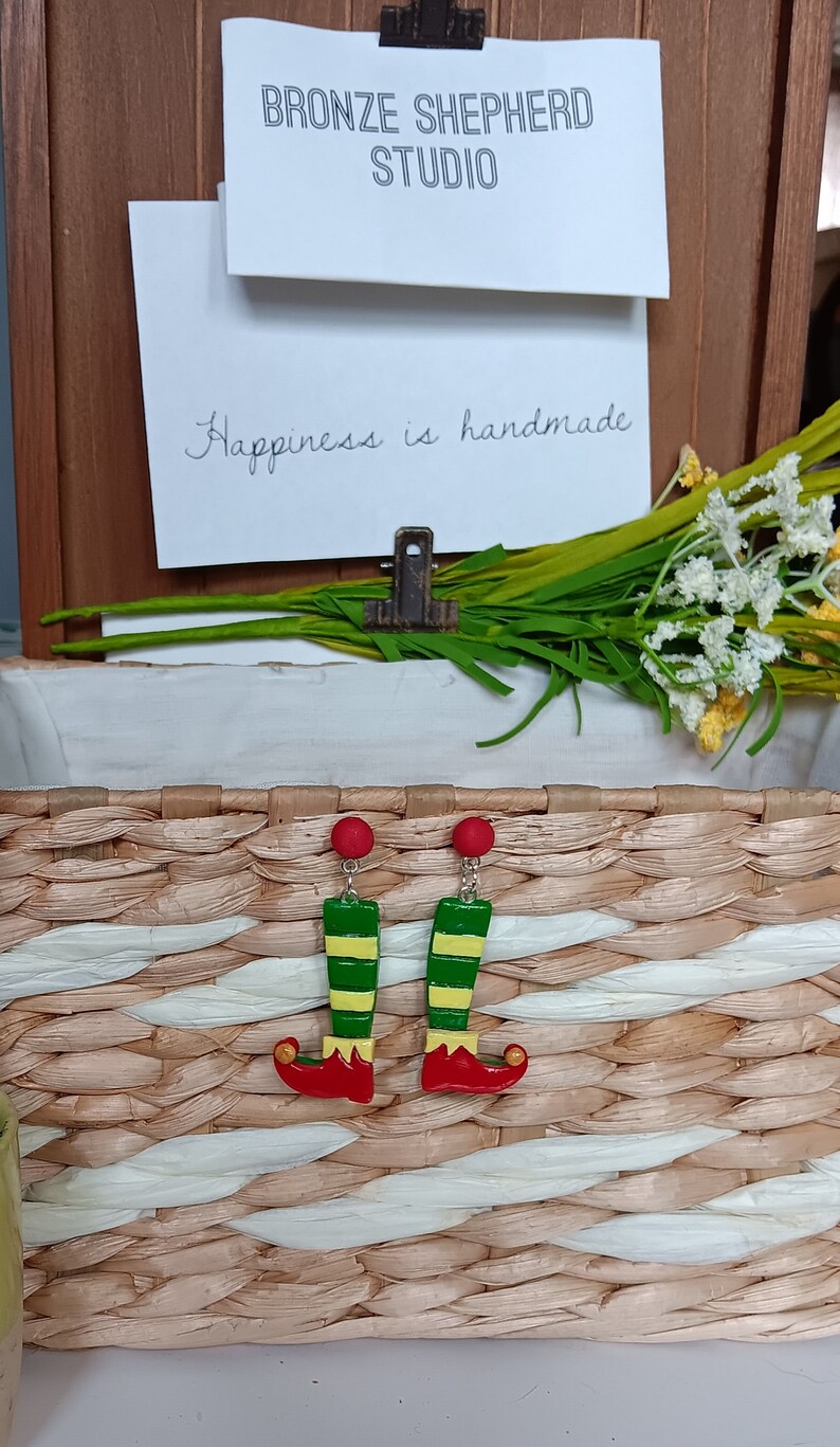 May include: A pair of elf leg earrings with red shoes, green and yellow striped legs, and gold bells. The earrings are displayed in a wicker basket.