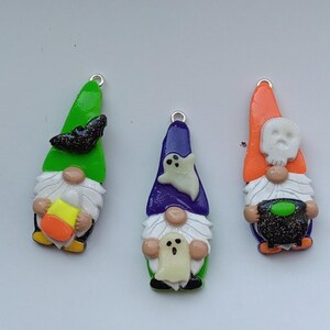 May include: Three Halloween gnome ornaments made of polymer clay. The gnomes are wearing green, purple, and orange hats. The gnome with the green hat has a black bat on his hat and is holding a candy corn. The gnome with the purple hat has a white ghost on his hat and is holding a white ghost. The gnome with the orange hat has a white skull on his hat and is holding a green cauldron.