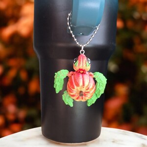 Könnte beinhalten: Ein schwarzer und grüner Kürbis-förmiger Schildkröten-Anhänger hängt an einer silbernen Kette. Der Anhänger ist an einem schwarzen und blauen Becher befestigt.