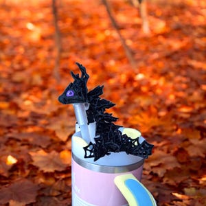 Pode incluir: Tampa de garrafa de dragão preto impressa em 3D com olhos roxos, sentada em cima de uma caneca de viagem rosa e amarela.