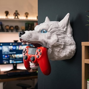 May include: A white wolf head wall mount sculpture with blue eyes and an open mouth holding a red video game controller. The wolf head is mounted on a dark wall, featuring detailed fur texture, a black nose, and visible teeth gripping the controller.
