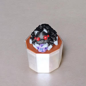 May include: A black and white spider figurine with red eyes sits on top of a white and brown octagonal dice tower. The tower has a purple 20-sided die inside.