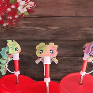 Pu&ograve; includere: Tre tappi per cannucce a forma di axolotl stampati in 3D e colorati. Ogni tappo &egrave; attaccato a una cannuccia rossa con un anello di plastica bianco. Le cannucce sono inserite in coperchi rossi con un anello di plastica bianco. I coperchi sono su tazze rosse.