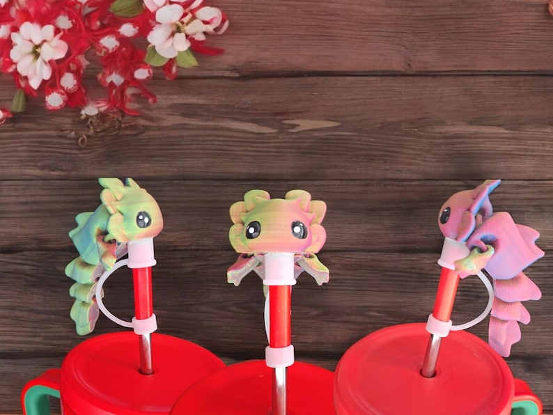 Pu&ograve; includere: Tre tappi per cannucce a forma di axolotl stampati in 3D e colorati. Ogni tappo &egrave; attaccato a una cannuccia rossa con un anello di plastica bianco. Le cannucce sono inserite in coperchi rossi con un anello di plastica bianco. I coperchi sono su tazze rosse.