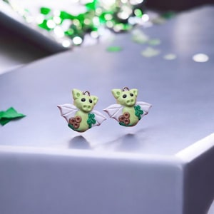 May include: A pair of light green dragon earrings with white wings and brown accents. The dragon earrings have a small gold loop at the top for hanging. The earrings are on a light gray surface.