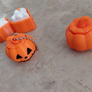 K&ouml;nnte beinhalten: Orangefarbene Halloween-Themen-Trinkets, darunter ein geschnitzter K&uuml;rbis mit einer silbernen Kette und ein separater k&uuml;rbisf&ouml;rmiger Beh&auml;lter. Au&szlig;erdem gibt es zwei orangefarbene und wei&szlig;e katzenf&ouml;rmige Gegenst&auml;nde. Die Gegenst&auml;nde befinden sich auf einer hellen Oberfl&auml;che.