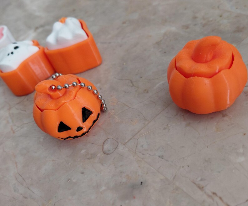 K&ouml;nnte beinhalten: Orangefarbene Halloween-Themen-Trinkets, darunter ein geschnitzter K&uuml;rbis mit einer silbernen Kette und ein separater k&uuml;rbisf&ouml;rmiger Beh&auml;lter. Au&szlig;erdem gibt es zwei orangefarbene und wei&szlig;e katzenf&ouml;rmige Gegenst&auml;nde. Die Gegenst&auml;nde befinden sich auf einer hellen Oberfl&auml;che.