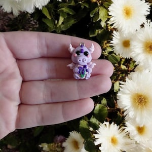 May include: A small, lavender-colored dragon figurine with white horns, wings, and a purple flower bouquet. The dragon has black eyes and is holding a bunch of purple flowers. The figurine is made of clay and has a silver loop at the top for hanging.