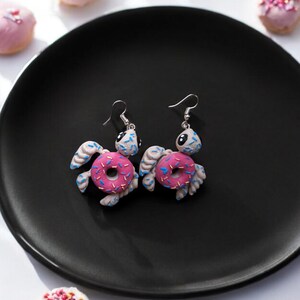 May include: A pair of silver dangle earrings featuring a white turtle with pink and blue donut accents. The turtle is holding the donut in its front flippers.
