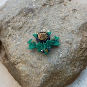 Puede incluir: Una figurita de dragón verde y dorada sosteniendo una olla de monedas de oro. El dragón tiene ojos negros y está sentado en una roca gris.