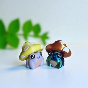 Op de afbeelding: Twee grillige kikkerbeeldjes met paddenstoelkap. Eén kikker is lavendelkleurig met een gele paddenstoelkap, de andere is blauwgroen met een bronzen kap. Beide hebben grote, expressieve ogen en staan op een witte achtergrond.