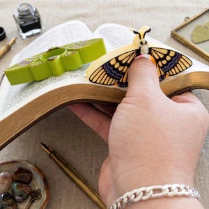 May include: An open book with gold-edged pages, featuring a yellow and black moth-shaped bookmark. A green ashtray and a small dish with stones and a key are also visible. A vintage inkwell and pen are on the table.
