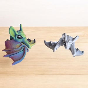 Puede incluir: Dos pequeñas figuritas de murciélagos, una es verde e iridiscente con alas moradas, la otra es gris con alas blancas.