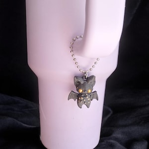 May include: A dark gray bat charm with yellow eyes and small wings hangs from a silver ball chain. The bat charm is attached to a light purple tumbler. The background is dark.