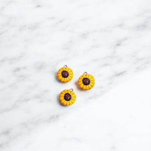 May include: Three yellow sunflower charms with brown centers. The charms have a silver loop for attaching to a necklace, bracelet, or other jewelry.