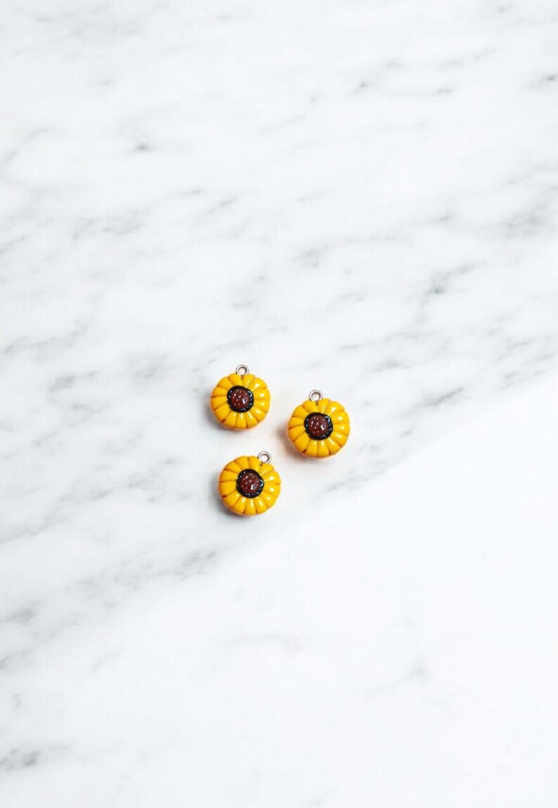 May include: Three yellow sunflower charms with brown centers. The charms have a silver loop for attaching to a necklace, bracelet, or other jewelry.