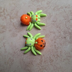 May include: Two green and orange Halloween spider charms with pumpkin accents. The spiders have purple dots on their bodies and are made of polymer clay.