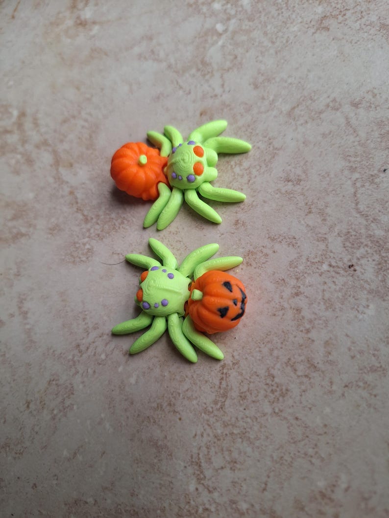May include: Two green and orange Halloween spider charms with pumpkin accents. The spiders have purple dots on their bodies and are made of polymer clay.