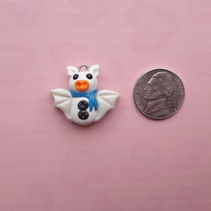 May include: A white bat-shaped charm with a blue scarf, orange nose, and black button details. A US nickel coin is next to the charm for size comparison. The charm has a silver loop for hanging.