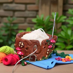 May include: A brown yarn bowl with a whimsical tree trunk design, featuring red and white mushroom accents. The bowl holds a ball of white yarn, with a crochet hook inserted. Colorful yarn skeins and a blue cloth are nearby.