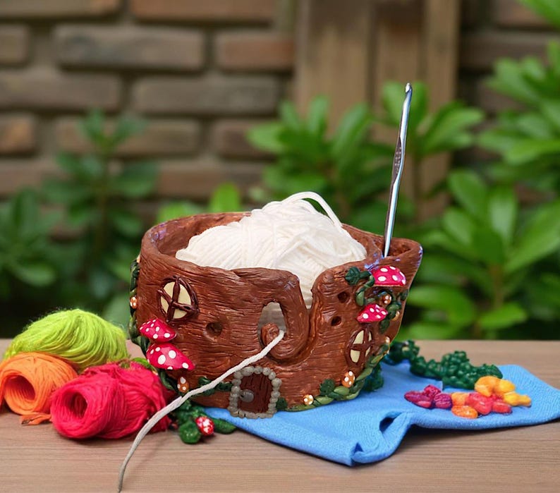 May include: A brown yarn bowl with a whimsical tree trunk design, featuring red and white mushroom accents. The bowl holds a ball of white yarn, with a crochet hook inserted. Colorful yarn skeins and a blue cloth are nearby.