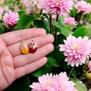 May include: Two small ceramic charms shaped like eggs with painted faces. One charm is yellow and the other is red.