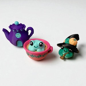Op de afbeelding: Een verzameling miniatuurkleifiguren. Een paarse theepot, een turquoise kikker in een roze kopje en een groene kikker met een heksenhoed en een pompoen.