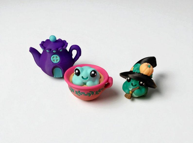 May include: A whimsical collection of miniature clay figurines. A purple teapot with a blue door, a teal frog in a pink teacup, and a green frog wearing a witch hat with a pumpkin.