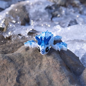 Può includere: Una piccola statuina di drago bianca e blu con occhi blu e una schiena spinosa, seduta su una roccia grigia.