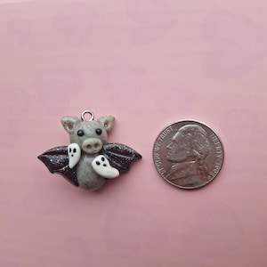 May include: A gray bat pendant with black glitter wings and two white ghost charms. The bat has a silver jump ring for hanging. A US quarter is next to the pendant for size comparison. The pendant is on a pink background.