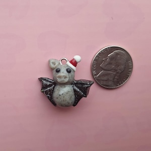 May include: A small, gray bat charm with black glitter wings and a red and white Santa hat. The bat is next to a U.S. nickel coin on a pink background. The charm has a loop for attaching to a necklace or bracelet.