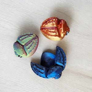 Puede incluir: Tres pequeñas figuritas de escarabajo de colores vivos. Una es azul, otra es verde y morada, y la última es naranja y dorada.