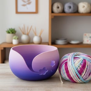 Puede incluir: Un cuenco de hilo morado y azul con un diseño floral, junto a una madeja de hilo colorido. El cuenco tiene una forma suave y curvada con detalles florales recortados. El hilo es una mezcla de rosa, azul y blanco.