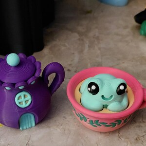 May include: A purple teapot with a light blue door and a matching lid, next to a pink teacup with a light blue frog figurine. A green frog wearing a witch hat is in the background. Whimsical, decorative items.
